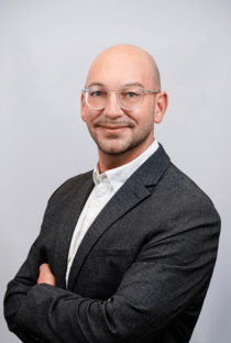 Porträtfoto eines Mannes mit Brille, rasiertem Kopf und dunklem Anzug.